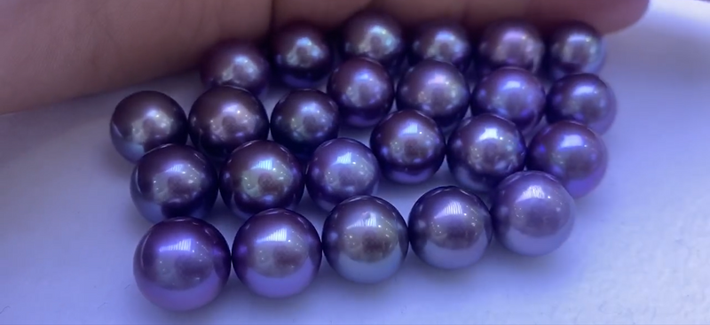 A pile of round, polished, dark purple pearls from Timeless Pearl with high luster scattered on a white surface.