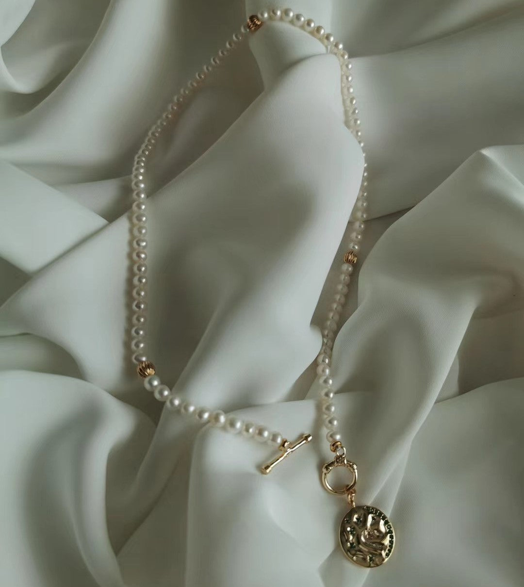 A delicate pearl necklace with a gold toggle clasp and coin pendant from Timeless Pearl, displayed on soft ivory fabric.