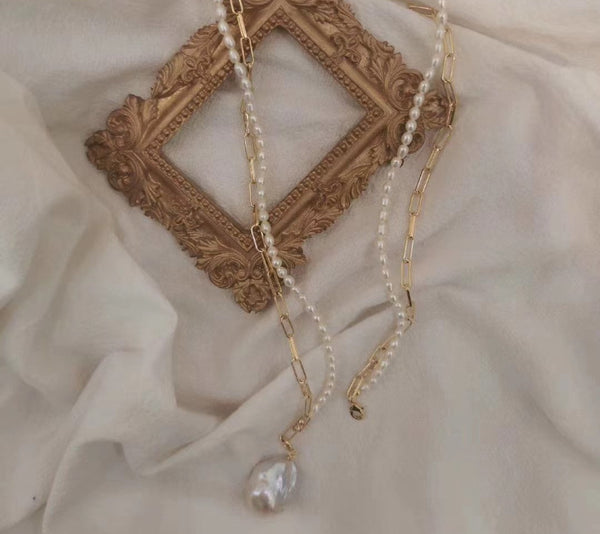 A gold and pearl necklace with a baroque pearl pendant from Timeless Pearl, displayed on a soft beige fabric with a vintage ornate frame.
