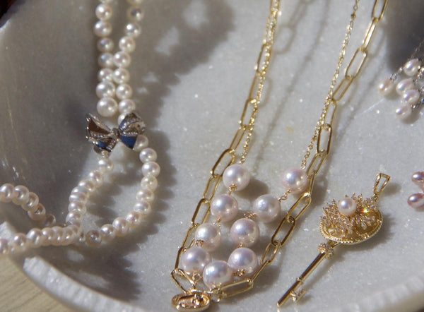 Timeless Pearl layered gold-chain necklaces and white pearl jewelry displayed in a ceramic dish under soft light.