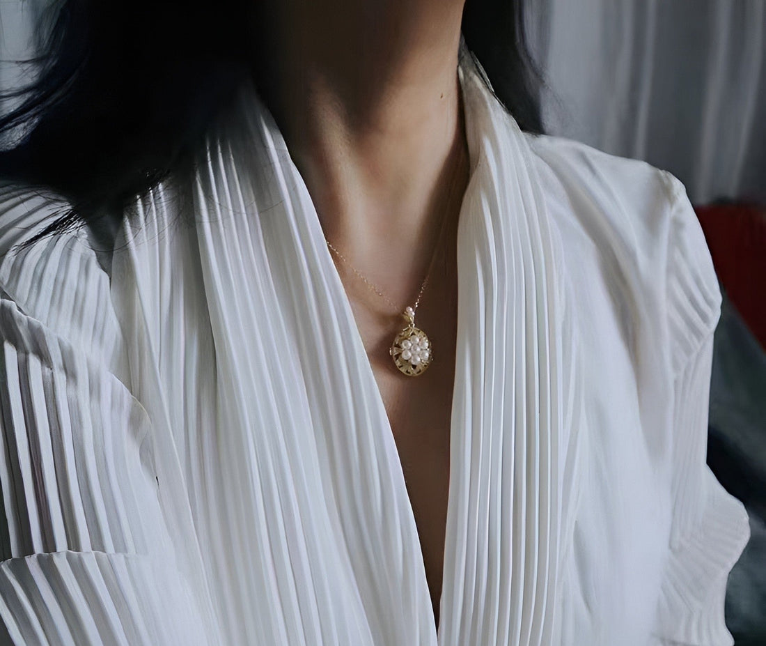 Woman wearing a white pleated blouse and a Timeless Pearl gold necklace with an ornate pearl drop pendant.